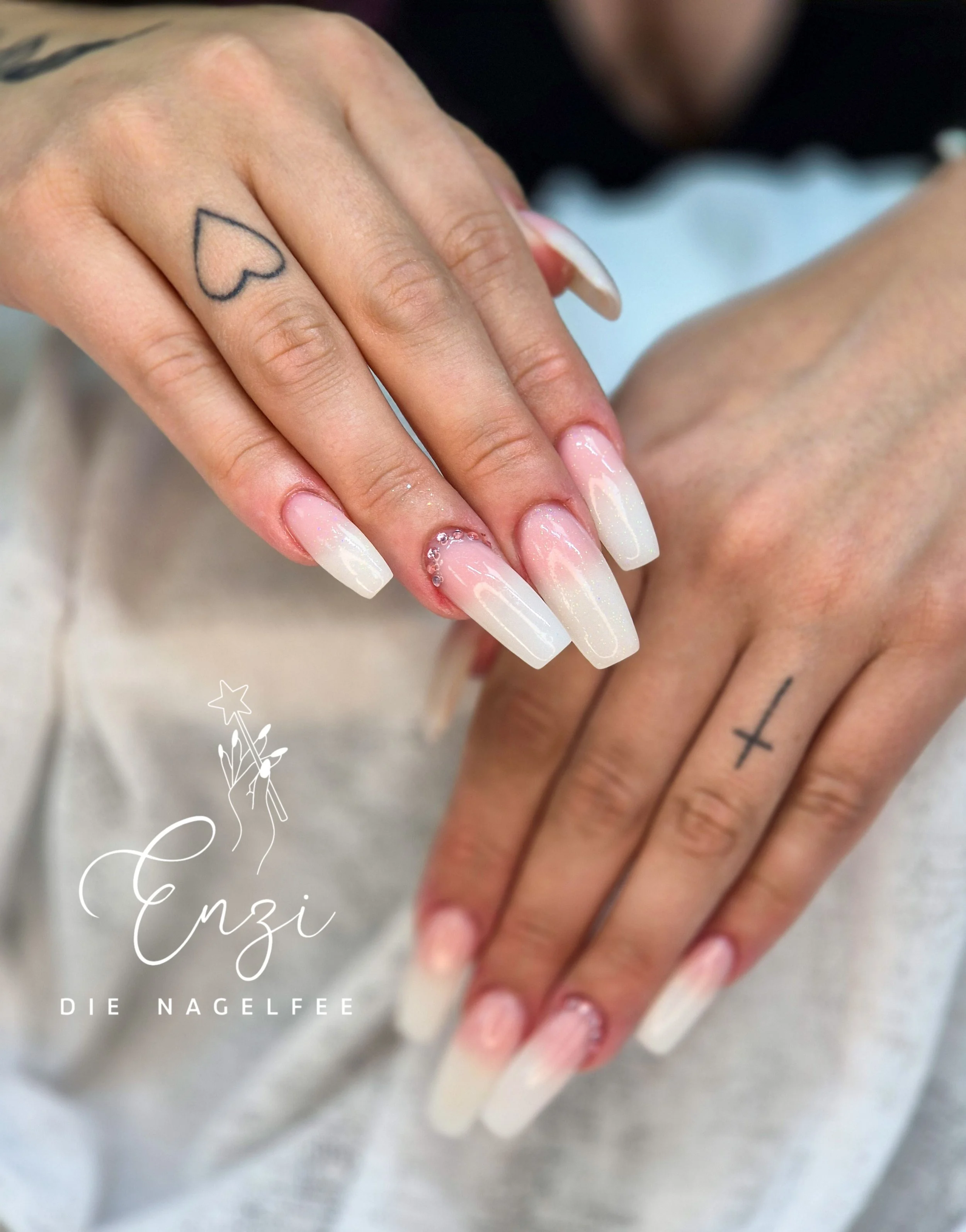 Nahaufnahme von Händen mit French Nails, einem Tattoo eines Herzens auf der linken Hand und einem Kreuz auf der rechten Hand, vor einem hellen Hintergrund.