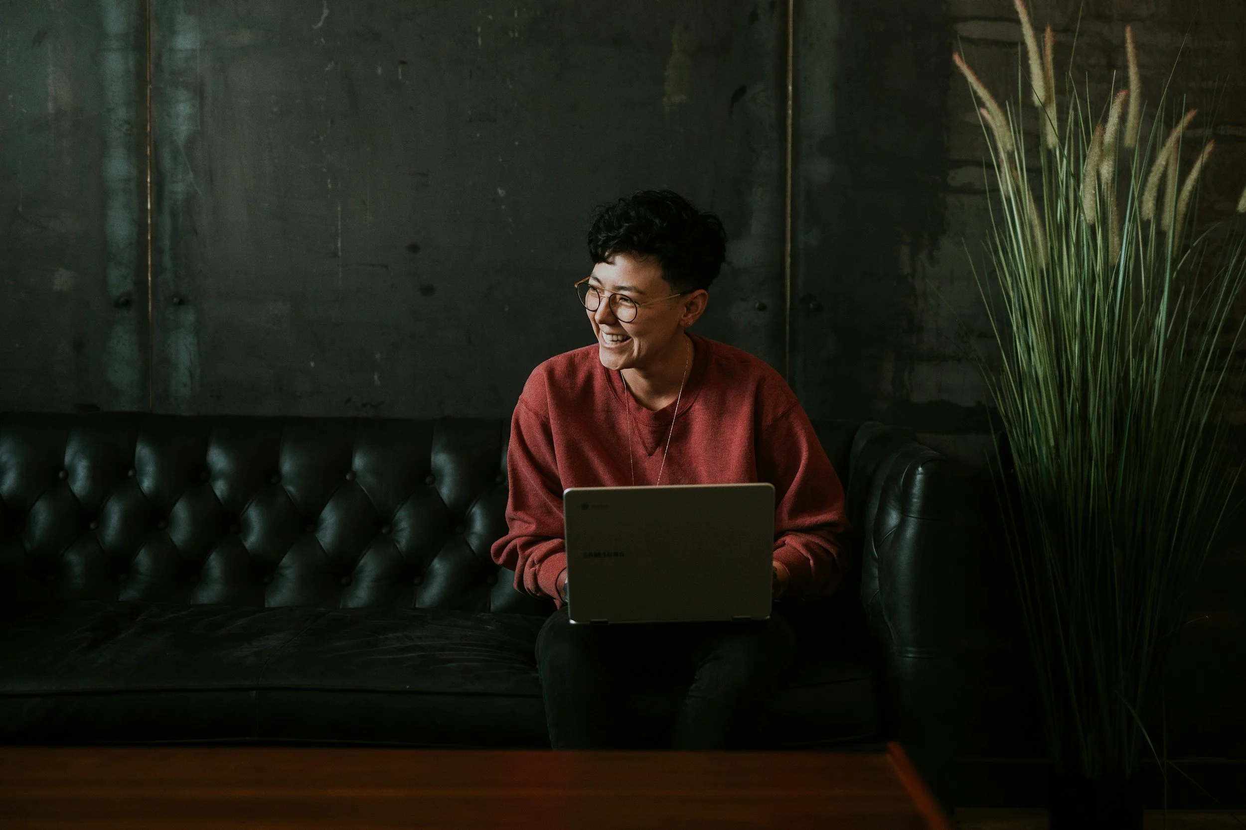 Person smiling while sitting on a black leather couch with a laptop on their lap, in a dimly lit room with a dark wall and a tall green plant.