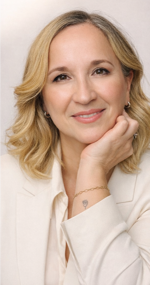 Mulher com cabelo loiro, sorriso suave, usando blazer branco, apoiando a cabeça na mão esquerda, com tatuagem de rosa no pulso, brincos pequenos e uma pulseira dourada.