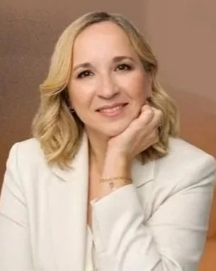 Mulher com cabelo loiro curvado, sorrindo, usando jaqueta branca, apoiando a mão no rosto