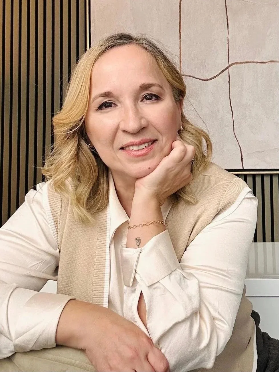 Mulher com cabelo loiro, vestindo camisa branca e colete bege, sentada em ambiente interno, sorrindo com o queixo apoiado na mão, em um fundo de parede de mármore e painéis de madeira.