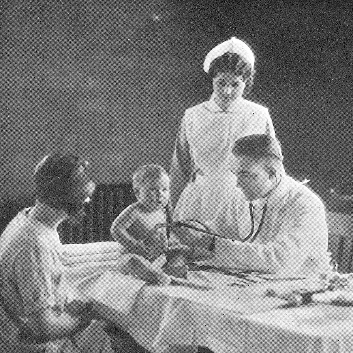 A vintage photo showing a doctor using a stethoscope to examine a baby sitting on a table, with a nurse and a woman, possibly the mother, observing.