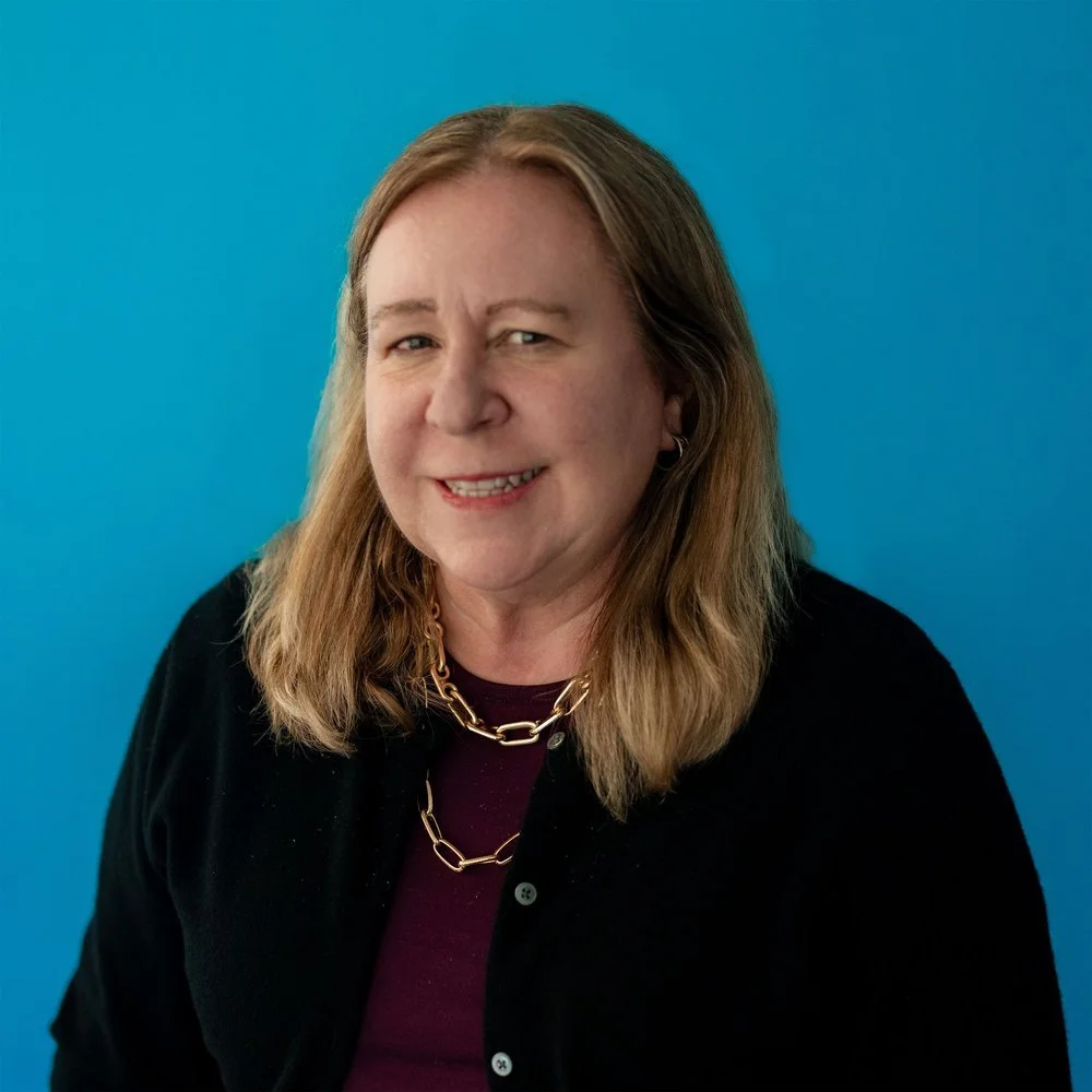 A woman with long hair wearing a black sweater and a gold chain necklace, standing against a blue background.