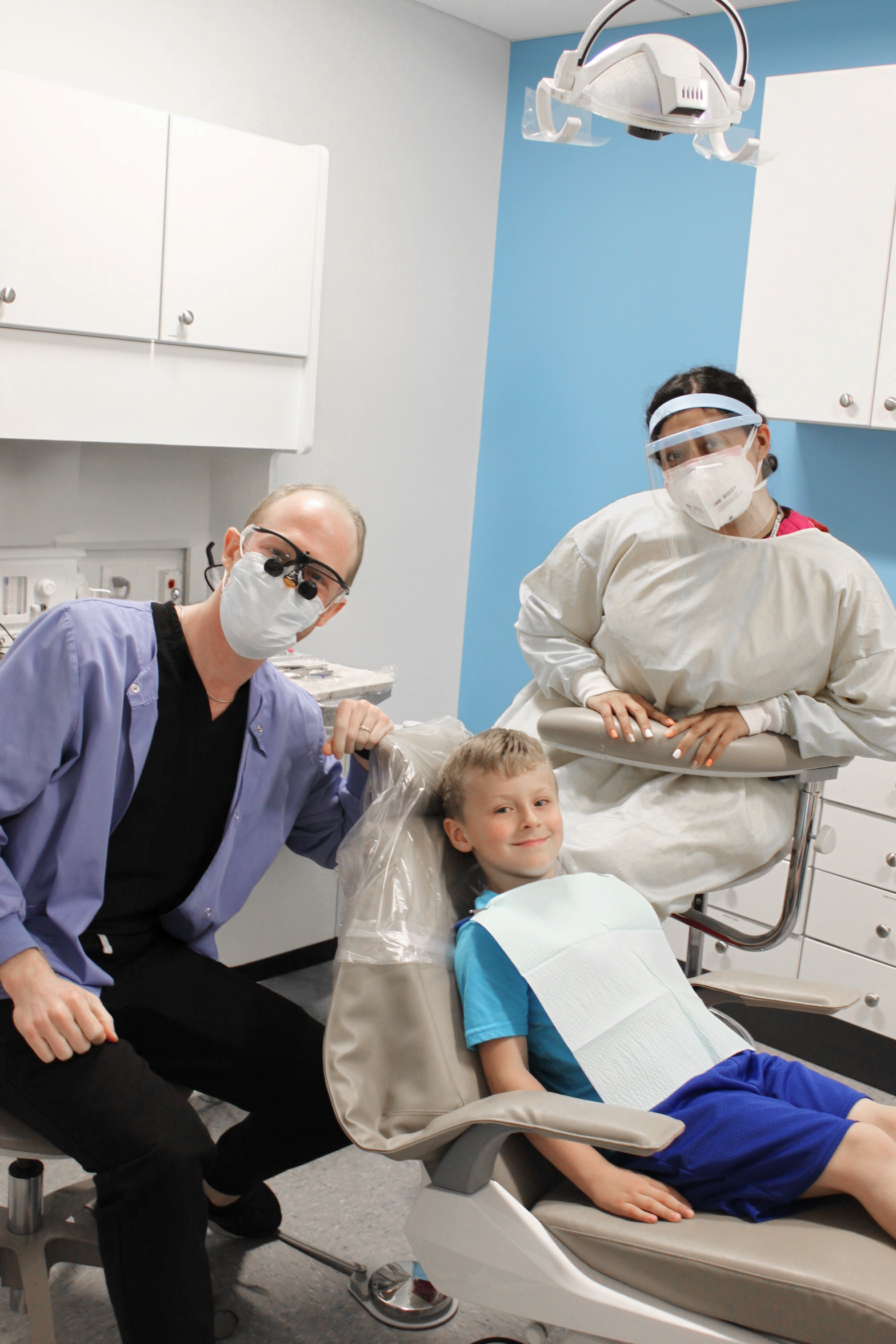 A child sitting in a dental chair, smiling, with a dental bib in front. A dentist wearing a mask and optical loupes is next to the child, along with a dental assistant wearing protective gear, including a face shield. The setting is a dental office.