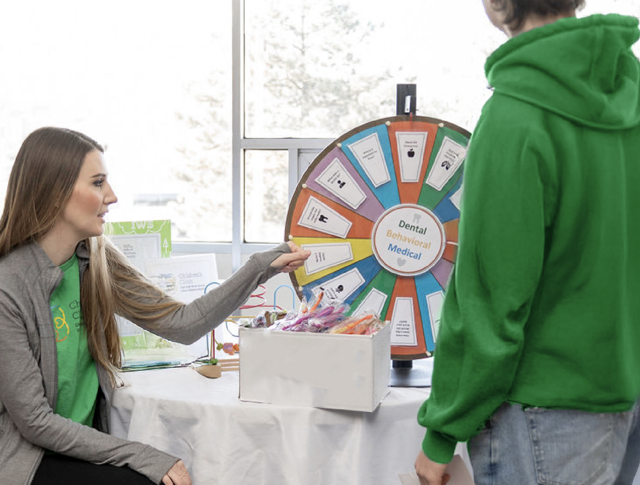 Two people at a table with a colorful prize wheel labeled "Dental Behavioral Medical" and a box of prizes.