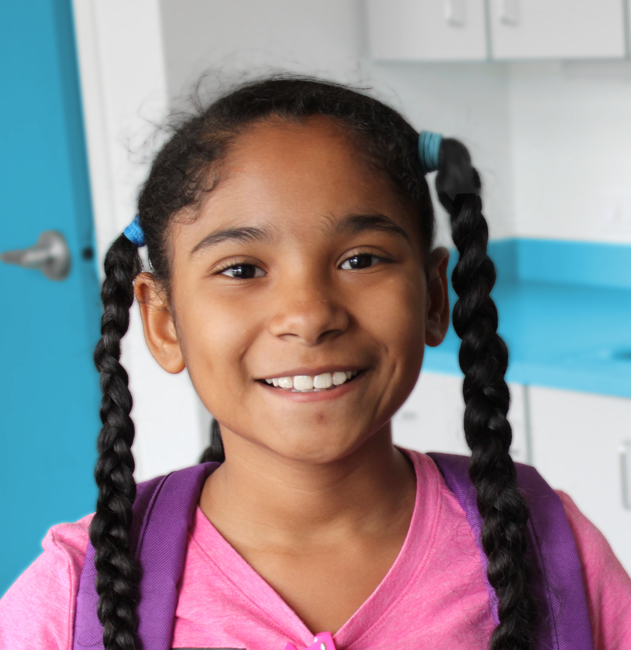 Smiling child with long braided hair wearing a pink shirt and purple backpack indoors.