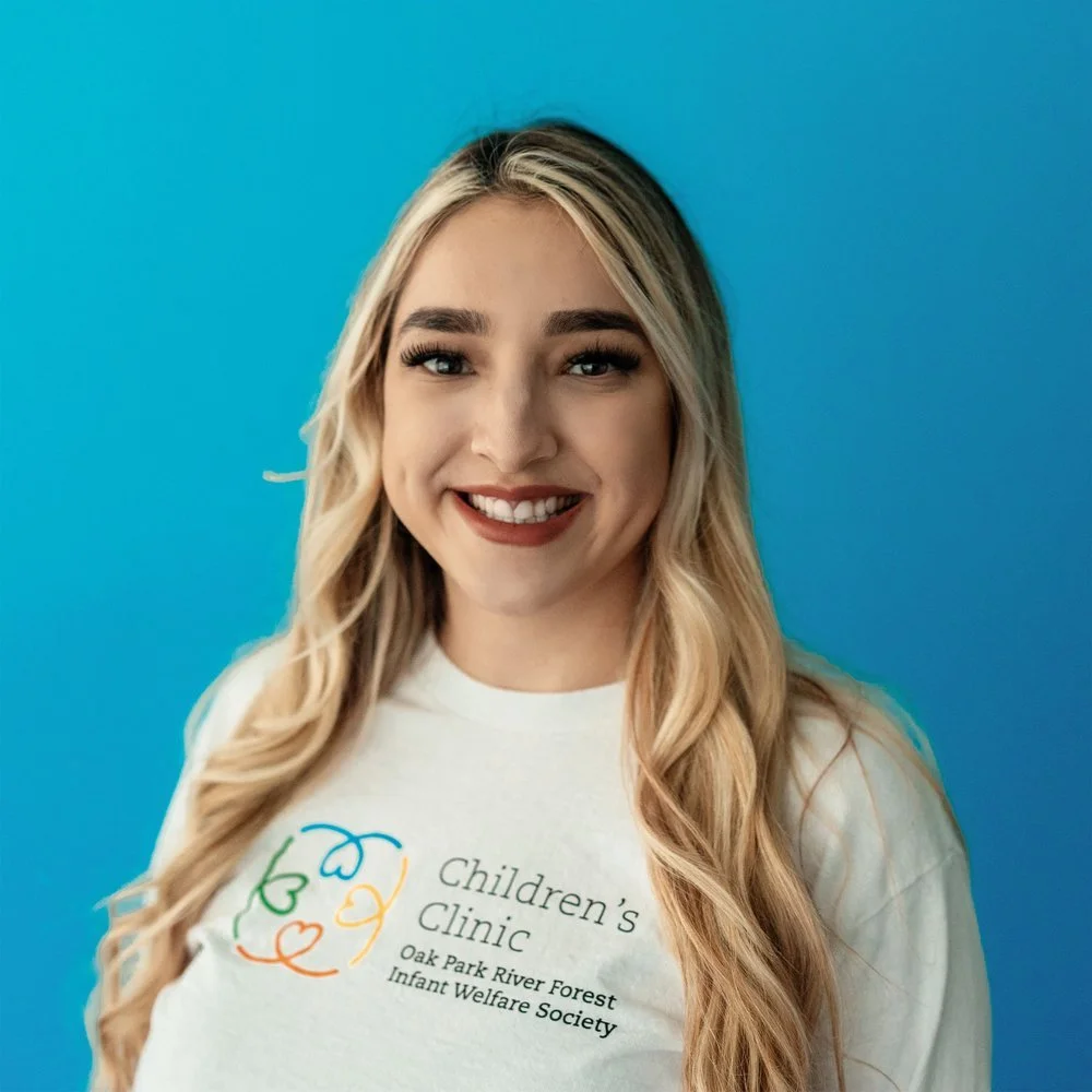 Woman smiling in front of a blue background, wearing a "Children's Clinic Oak Park River Forest Infant Welfare Society" t-shirt.