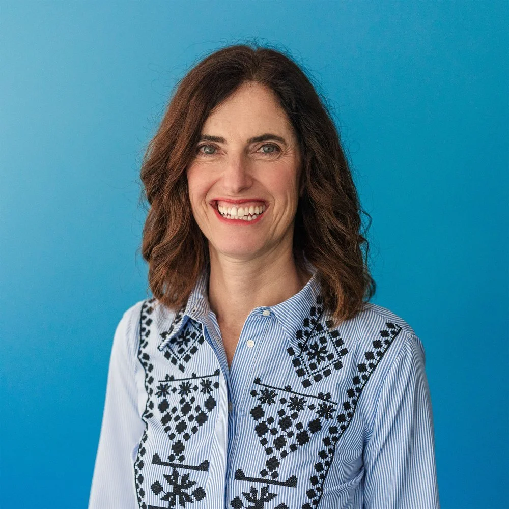 Smiling woman with curly hair in a patterned shirt against blue background.