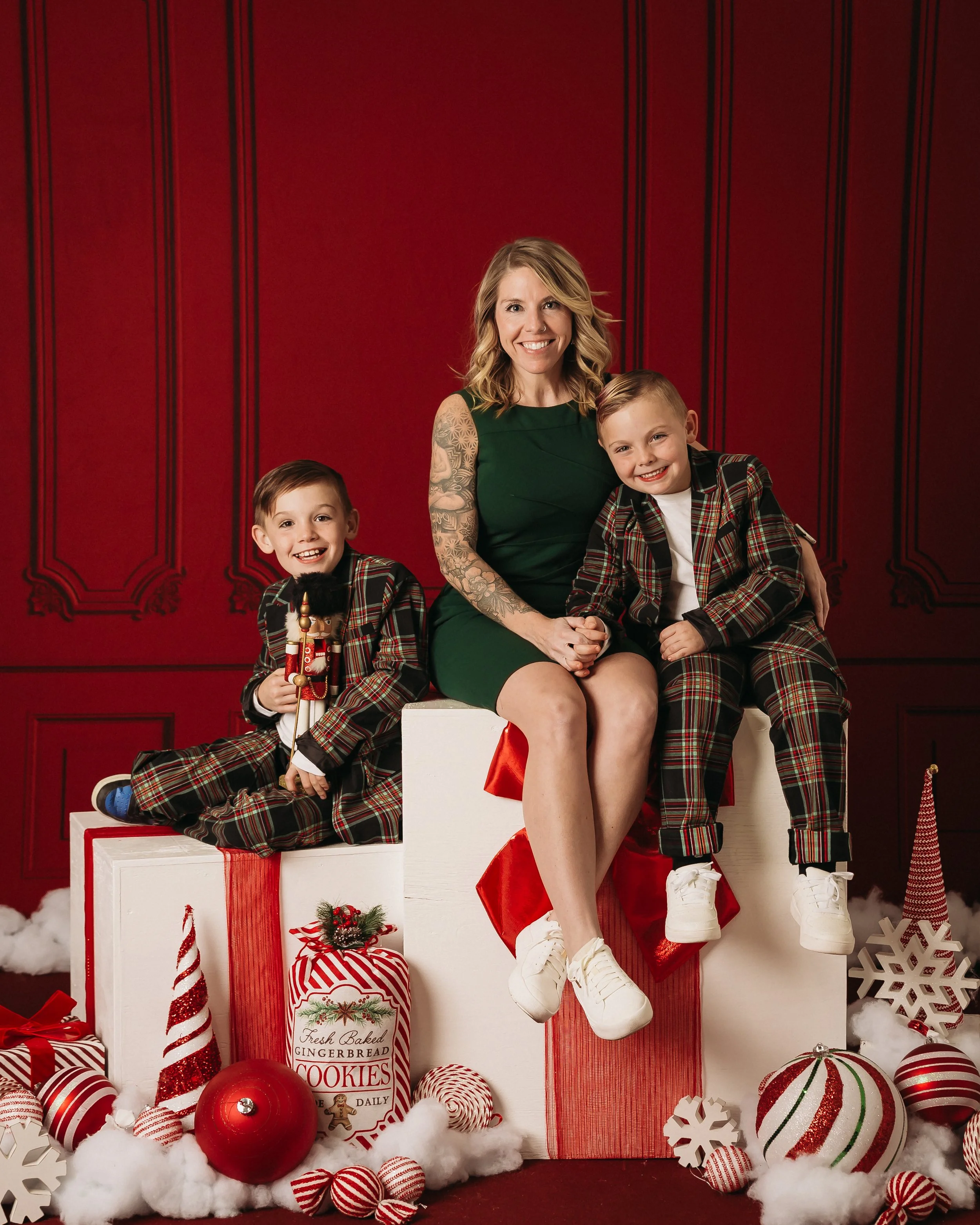 A family Christmas portrait featuring a woman in a green dress sitting between two boys in plaid suits. They are seated on large white gift boxes with red ribbons, surrounded by festive decorations like candy canes, snowflakes, and Christmas ornaments. The background is a rich red with decorative trim.