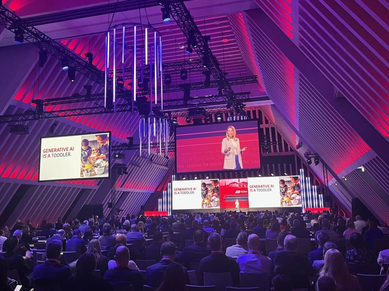 Conference with speaker discussing generative AI, large audience, modern auditorium with red and blue lighting.
