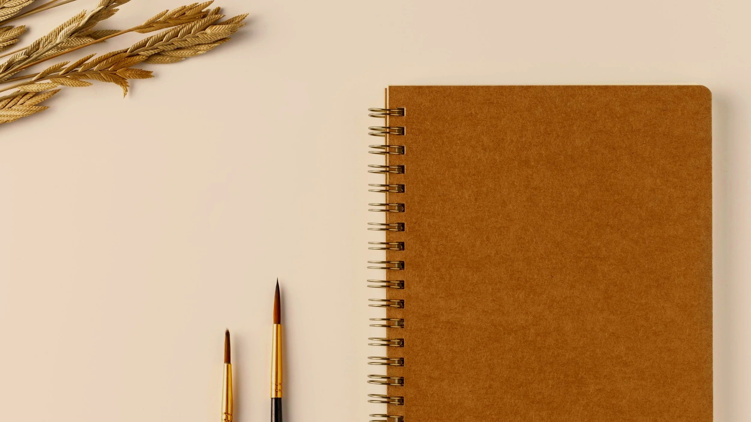 Flat lay of a brown spiral notebook, two paintbrushes, and dried wheat stalks on a white surface.
