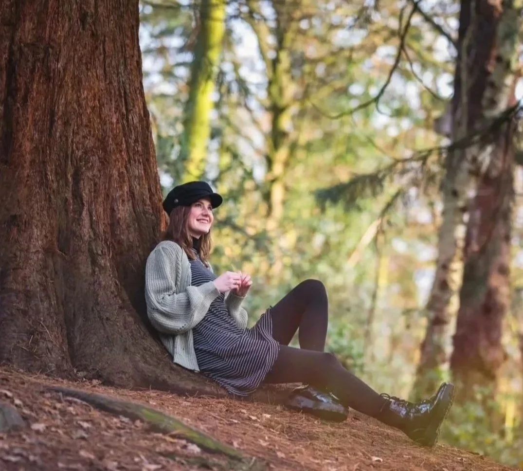 A woman sits on the ground and leans against a large tree trunk in a forest, smiling and looking upward. She is wearing a black hat, grey cardigan, striped dress, black tights, and boots, surrounded by tall trees with sunlight filtering through the leaves.
