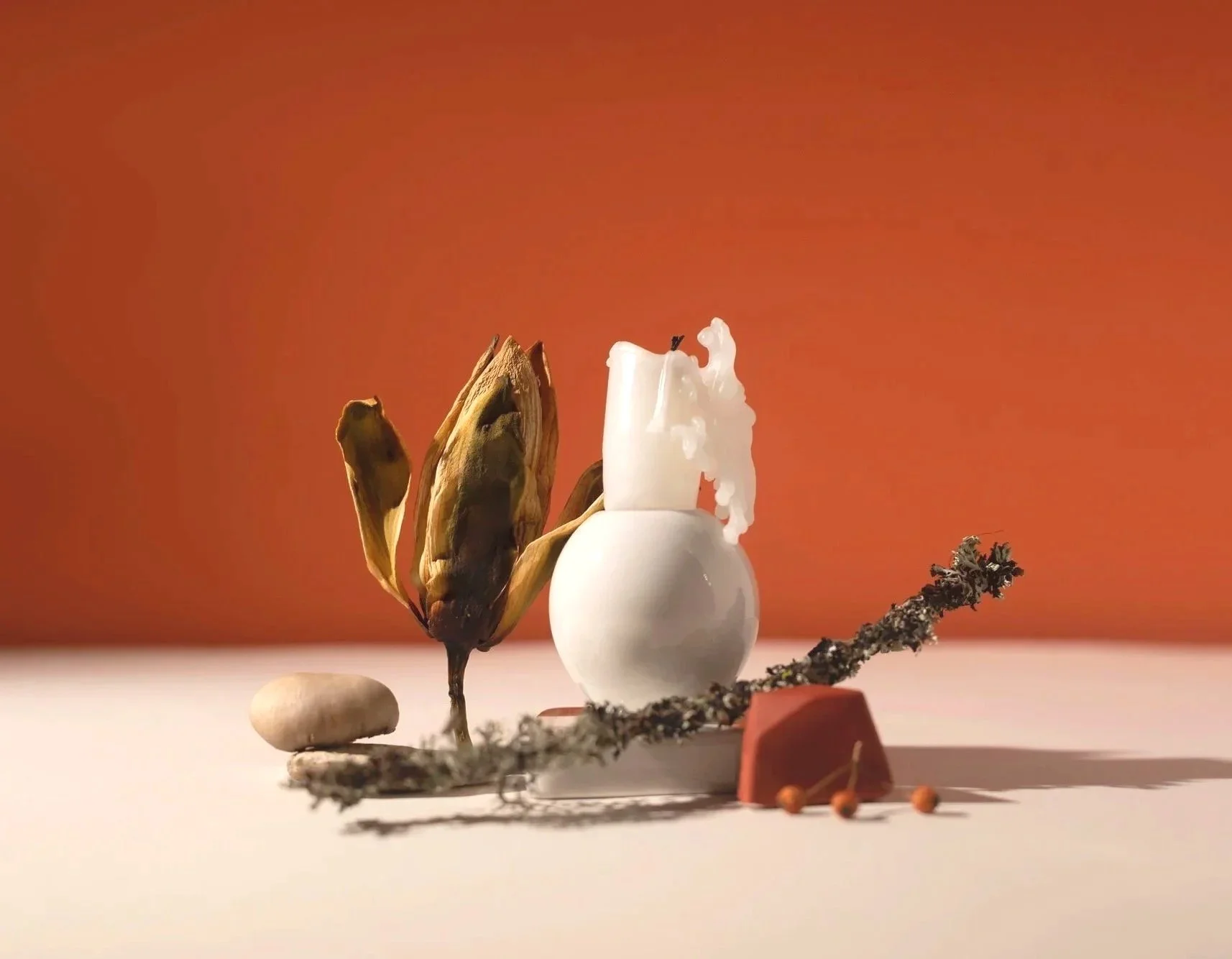 Still life arrangement with a dried banana peel, a sprig of dried moss, a smooth pebble, an egg-shaped white ceramic vessel with a melting candle, a red geometric block, and small orange berries on a white surface against an orange background.
