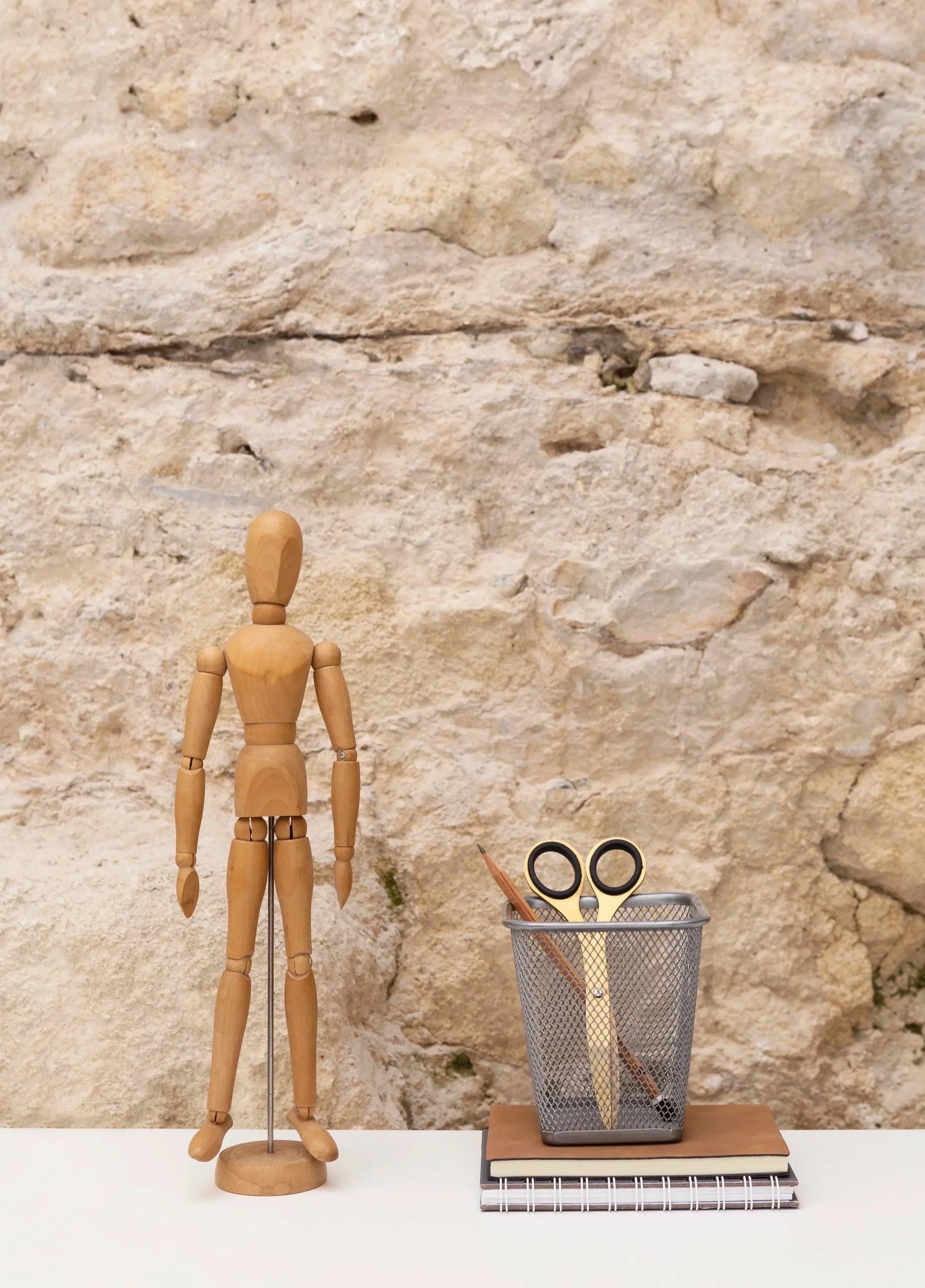 A wooden artist mannequin, a metal mesh pencil cup with scissors and a pencil, and stacked notebooks on a white surface in front of a stone wall background.