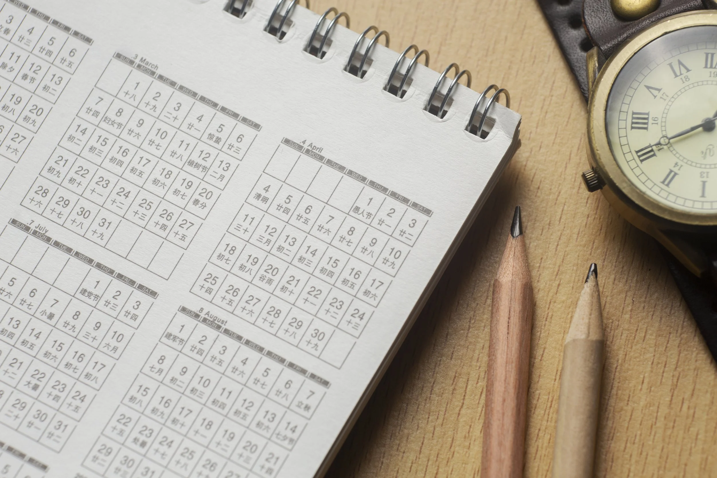A tabletop with a spiral-bound calendar showing months with dates and Chinese characters, two wooden pencils, and a round watch with a leather strap.