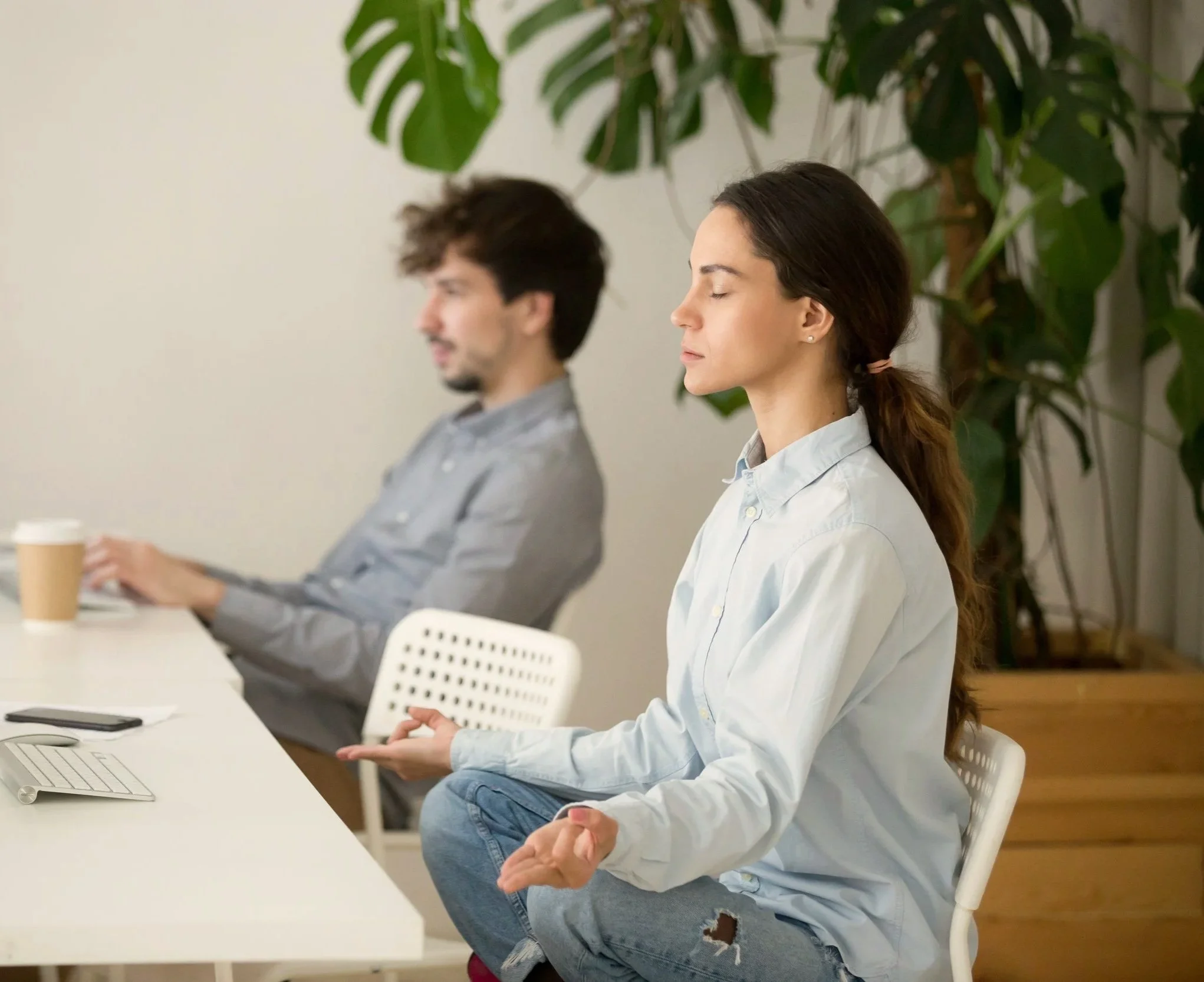 A woman with long brown hair tied back in a ponytail and a light blue shirt practicing meditation in a modern office, sitting cross-legged with eyes closed. In the background, a man with curly hair and a gray shirt, sitting at a desk with a coffee cup and a phone, also has eyes closed, with a large green plant behind them.