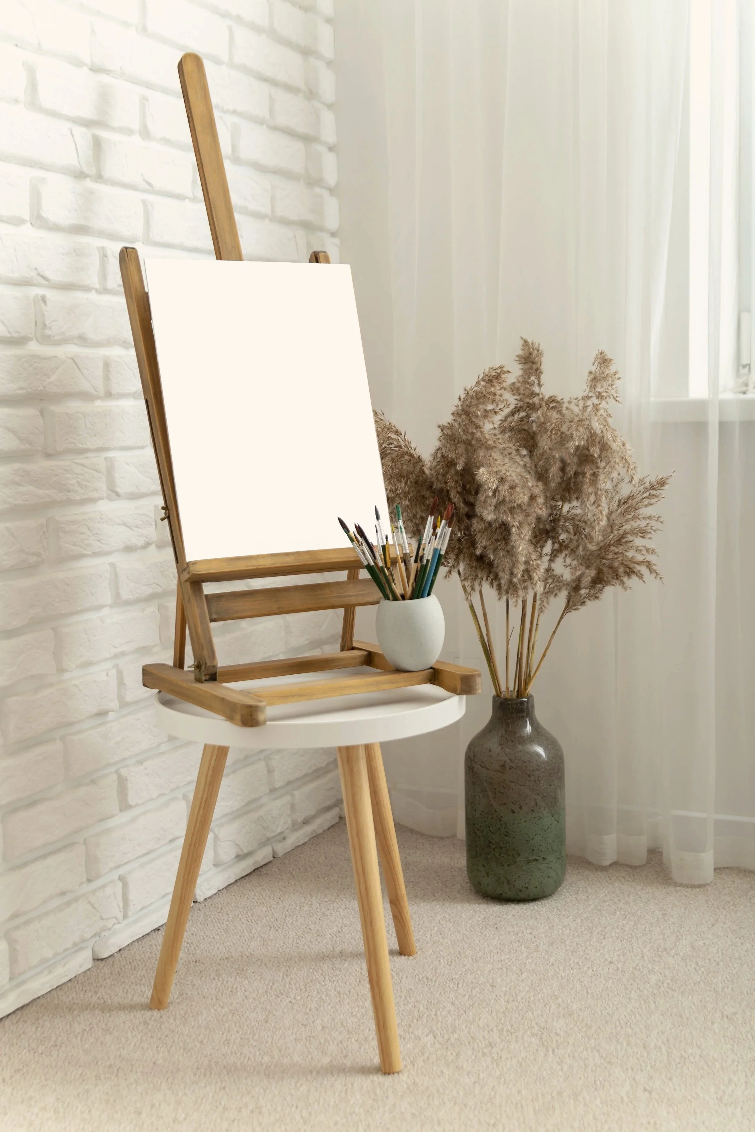 An art easel with a blank canvas, a vase with coloured pencils, and a large vase with dried pampas grass, set against a white brick wall and sheer curtains. A scene for private art therapy.