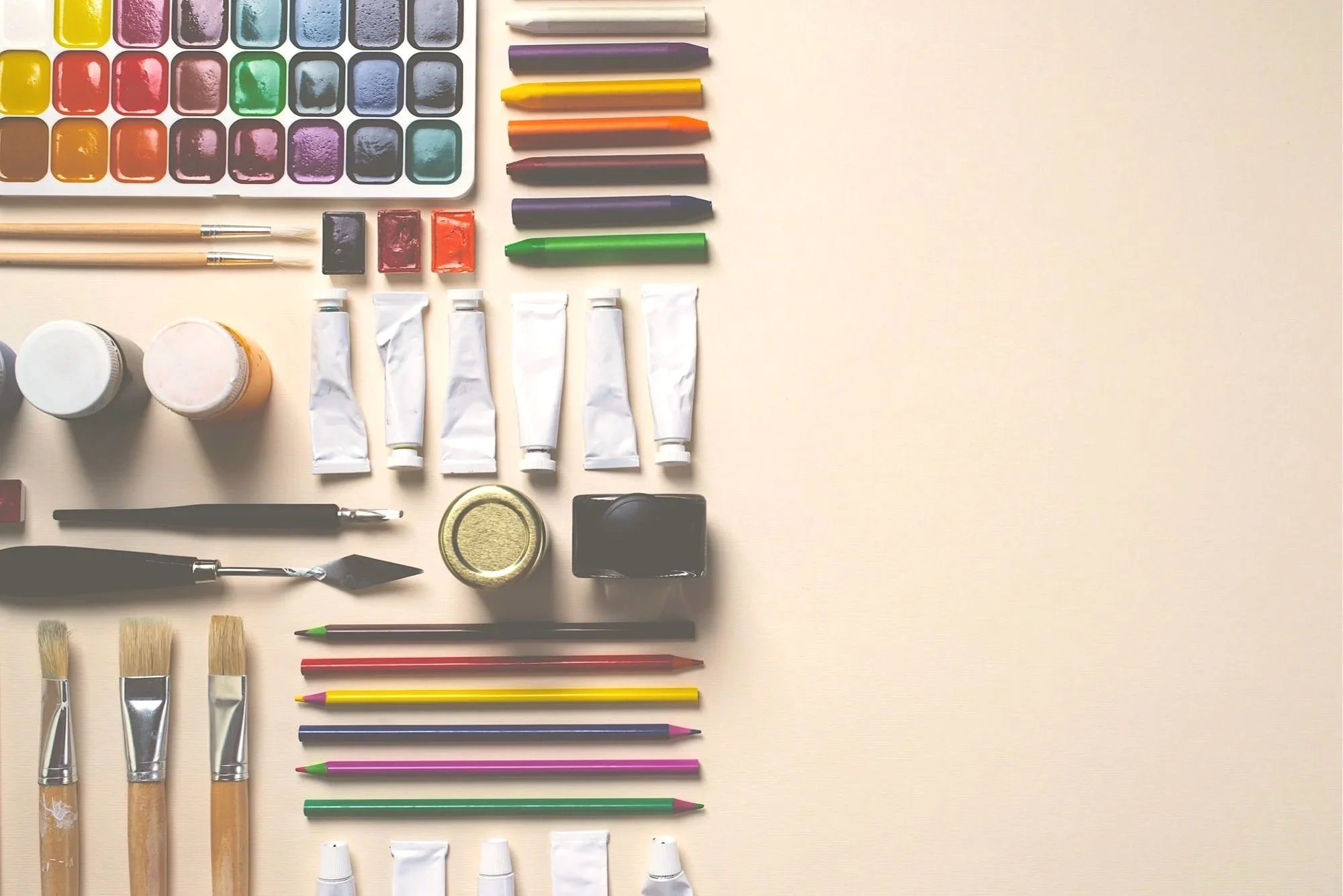 Art supplies arranged on a light-colored surface, including watercolor paints, paintbrushes, colored pencils, paint tubes, and small containers.