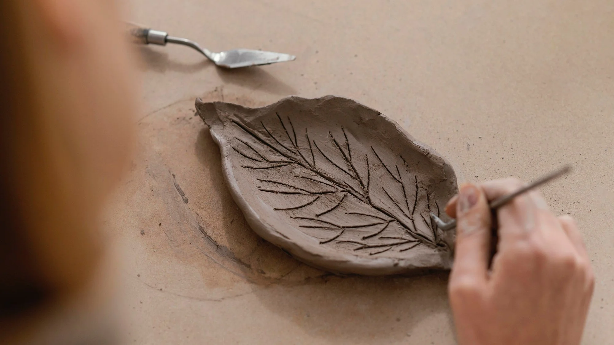Person carving a leaf pattern into a clay bowl with a small tool, with additional tools visible nearby.