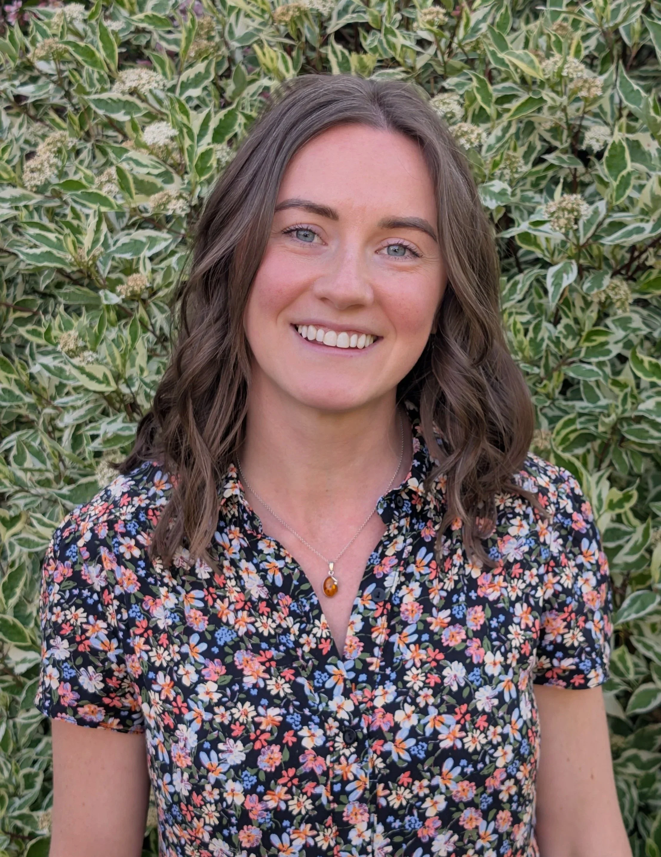 A young woman with shoulder-length wavy brown hair, blue eyes, and a friendly smile, standing outdoors in front of green variegated foliage, wearing a floral blouse and a necklace with a amber-colored pendant.
