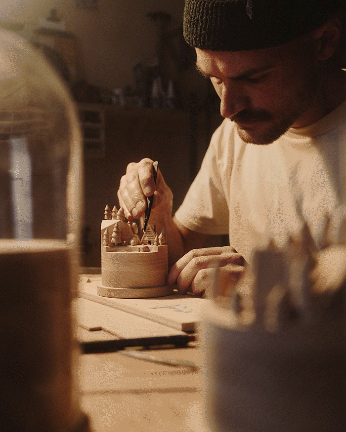 L'artisan Thibaut Malet travaillant sur une sculpture en bois miniature avec détails de petites maisons et arbres.