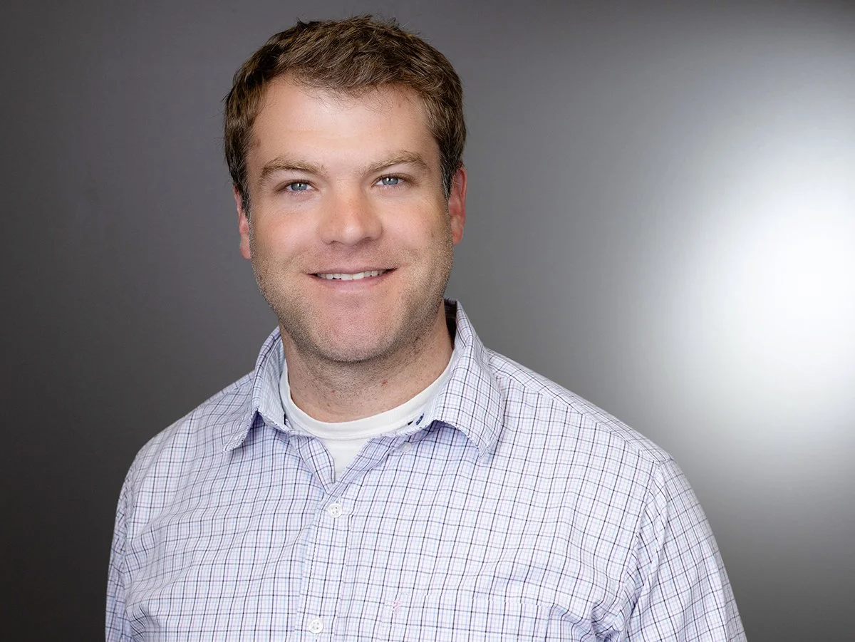 Rian Shimek, Portrait of a smiling man with brown hair wearing a light-colored plaid shirt against a gray background.