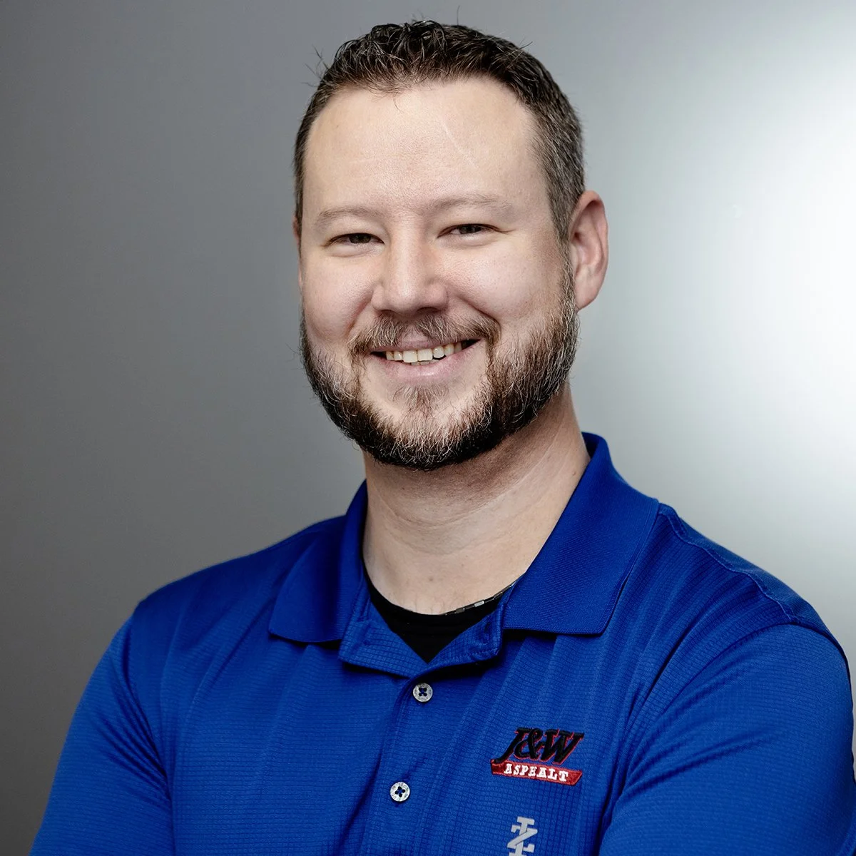 Kevin Kalina Headshot of a man with a beard smiling, wearing a blue polo shirt with patches, against a gray background.