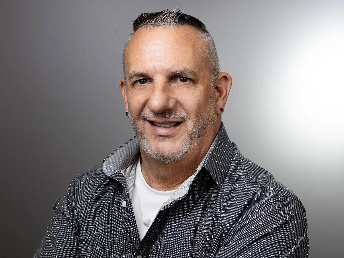Lon Ross, A smiling middle-aged man with short gray hair, a beard, and earrings wearing a black and white polka dot shirt against a gray background.