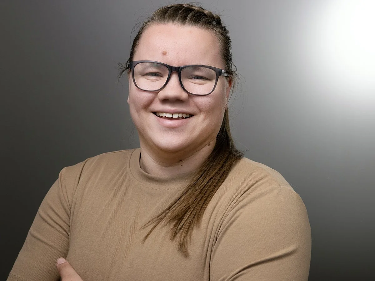 Katie Foley, A smiling person with glasses, long brown hair, wearing a tan shirt, crossed arms, standing against a gray gradient background.