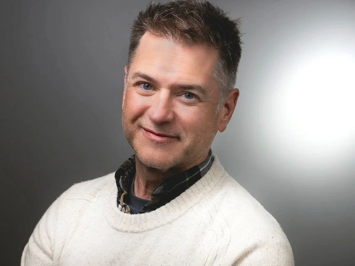 Tate Carlson, Marketing Integration Manager, man with short, dark hair and blue eyes, smiling slightly, wearing a cream sweater over a checkered shirt, against a gray background with a light source on the right.