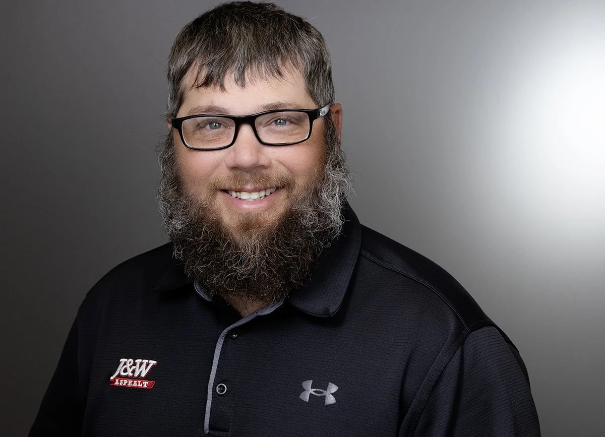 A smiling man with glasses and a beard, wearing a black Under Armour polo shirt with an J&W Asphalt logo, against a gray background.