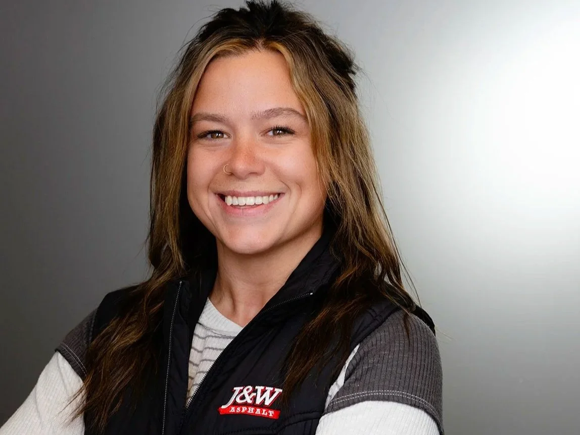 Angela Horski, A woman with long light brown hair smiling, wearing a black vest with a J&W Asphalt logo over a white shirt, against a neutral background.