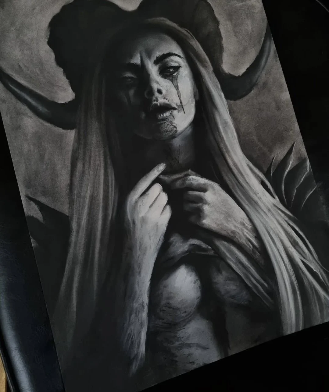 Monochrome drawing of a female figure with horns, tears and blood on face, long hair, and hands near chest, dark background.