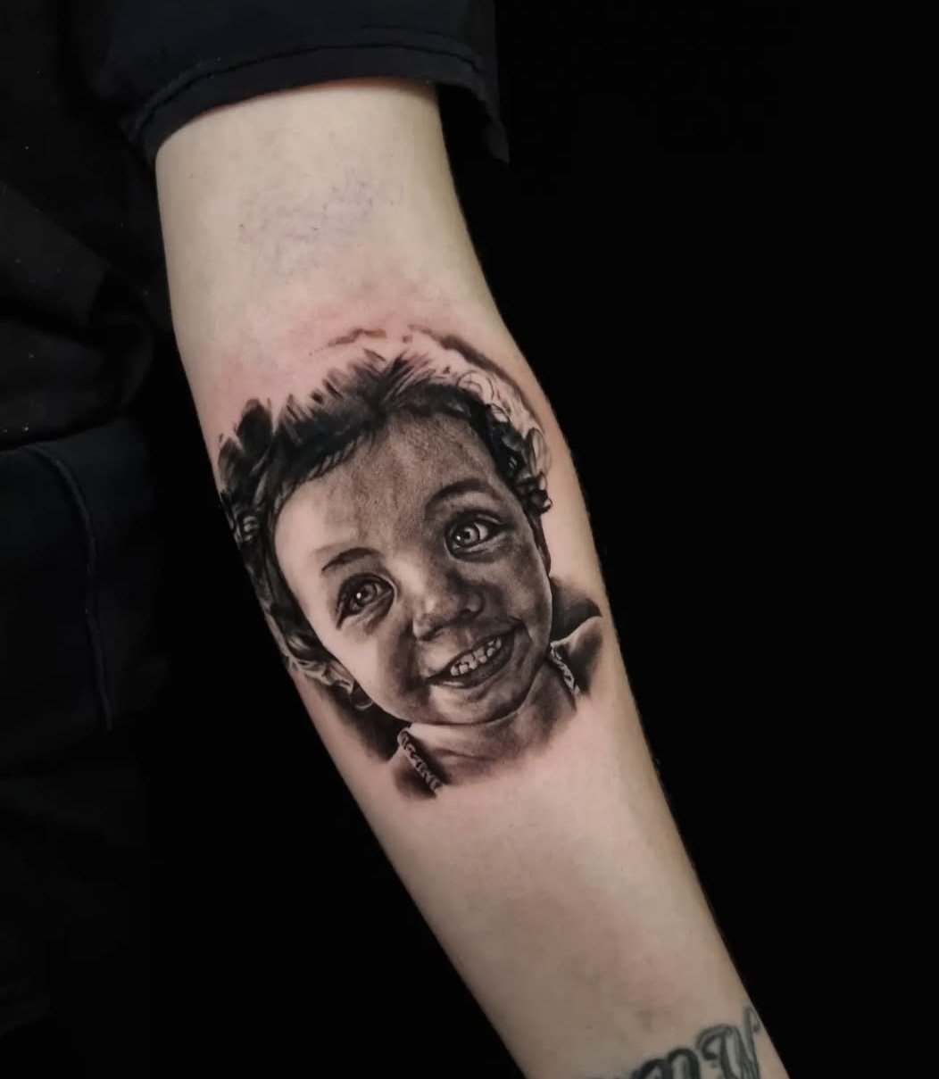 Black and white portrait tattoo of a smiling child's face on a person's forearm.