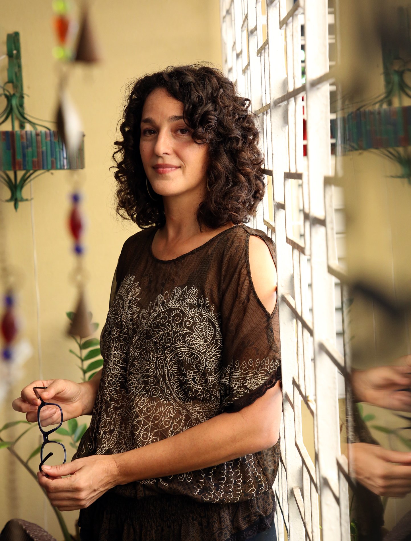 Mujer con cabello rizado y gafas en la mano, vestida con blusa negra bordada, frente a una ventana de diseño geométrico.