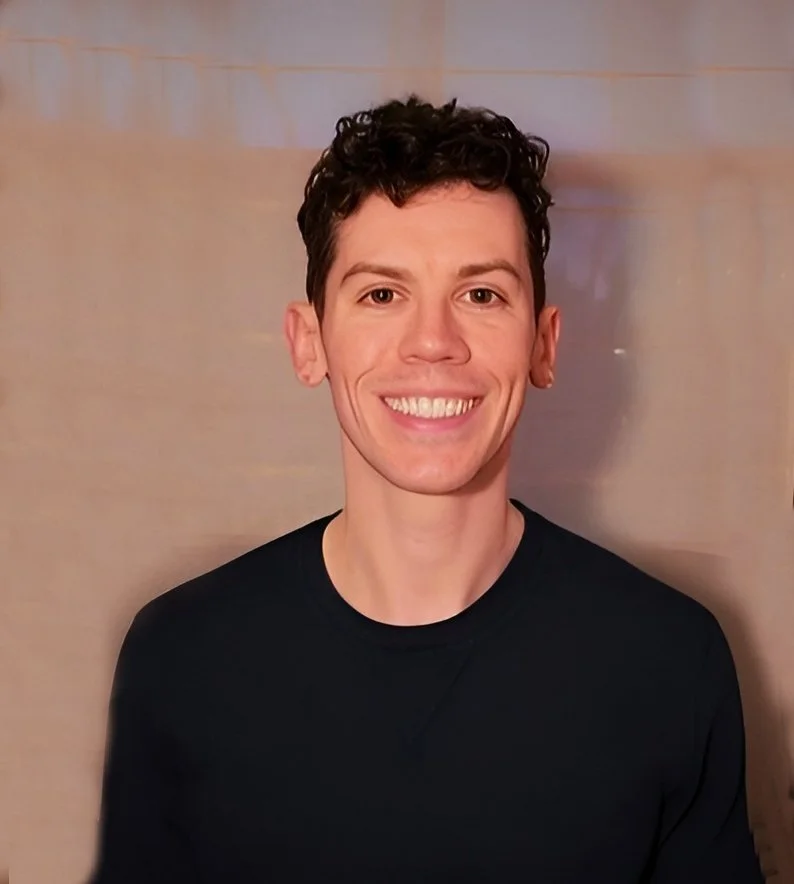 A young man with curly dark hair, smiling, wearing a black shirt, standing in front of a neutral background.