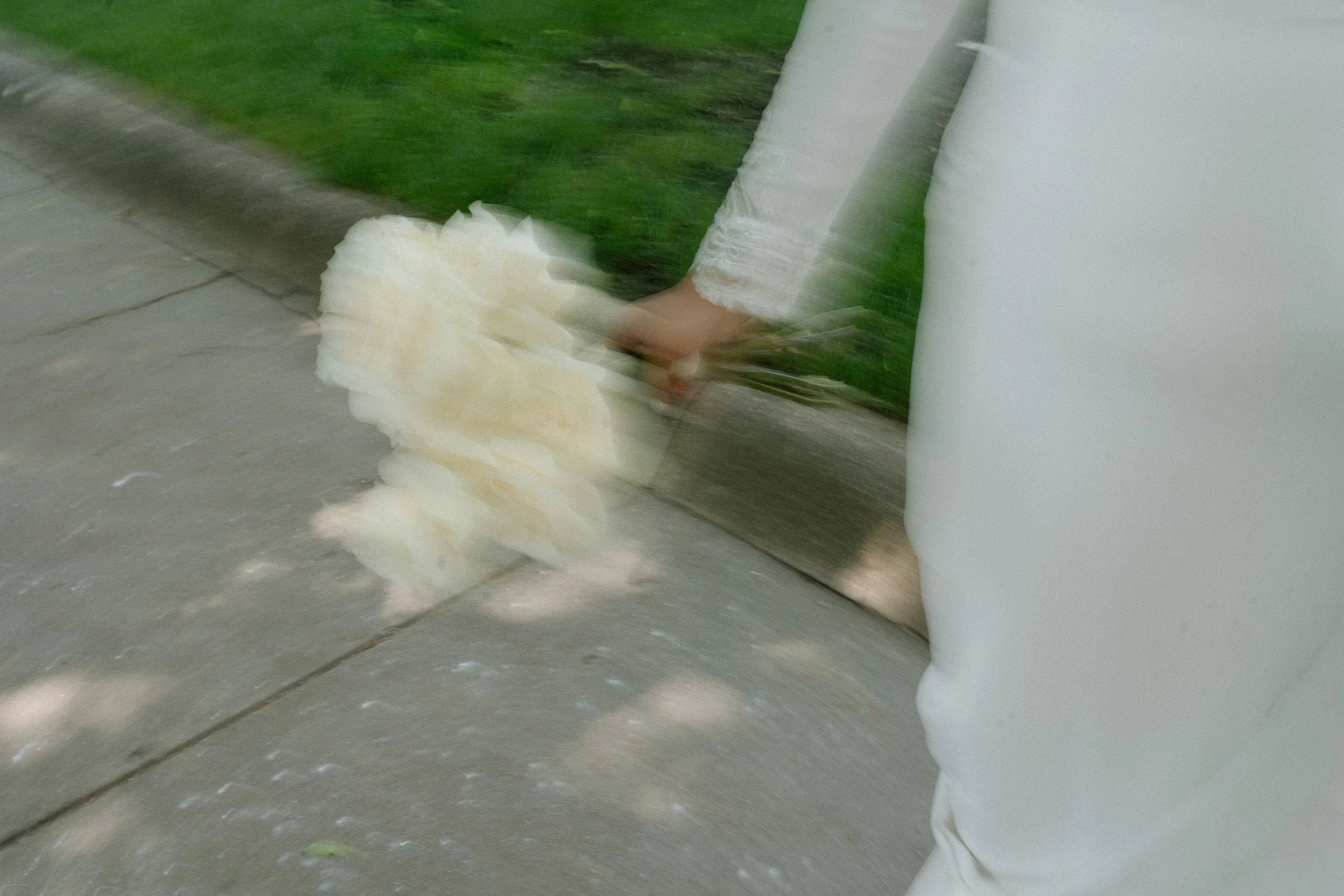 A person's foot stepping on a white flower laying on the ground next to a building wall, with a patch of grass nearby.
