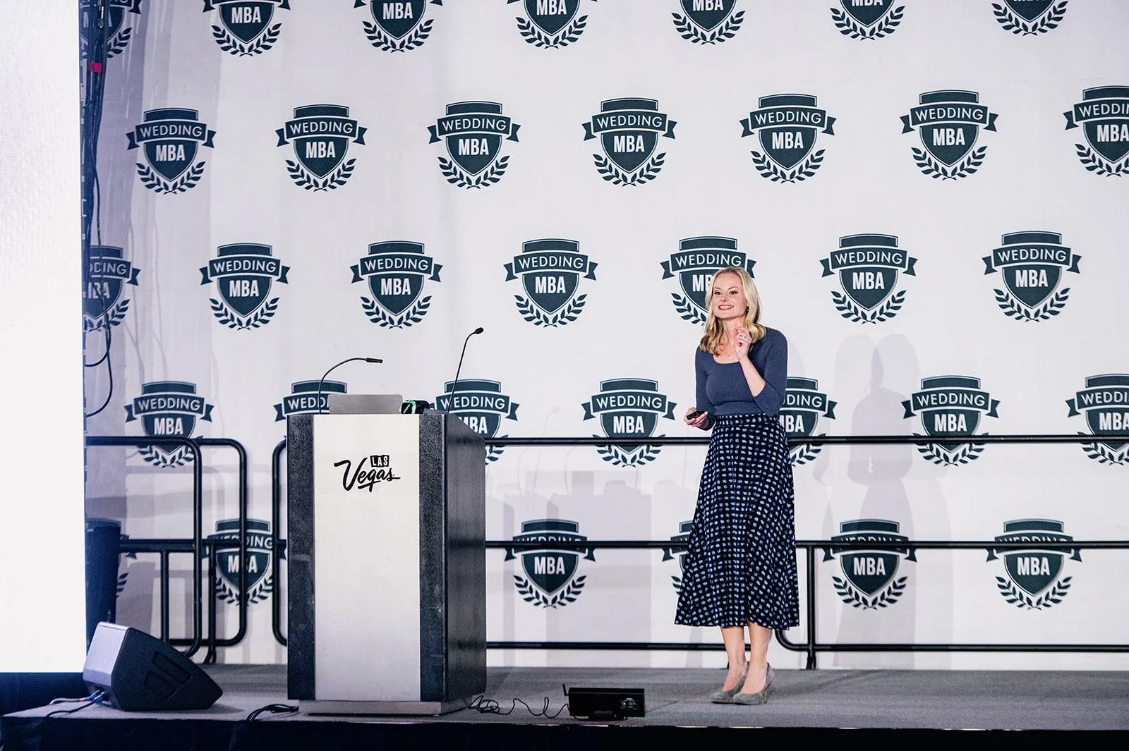A woman stands on a stage at a conference, with a backdrop featuring multiple logos reading 'Wedding MBA.' She is holding a presentation remote and smiling, wearing a navy top and a long, polka dot skirt. A podium with the word 'Las Vegas' is nearby, and there are microphones and audio equipment on the stage.