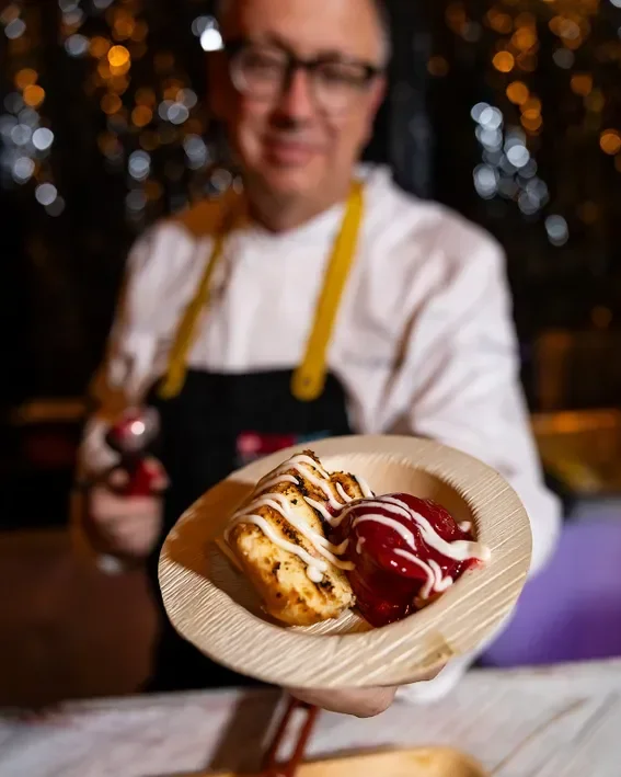 Entrance to a bar area with a neon sign that reads 'Oh how sweet it is'. The bar has purple and gold decor, with plants and white decorative panels on either side. There are red carpets and gold stanchions at the entrance to the bar.