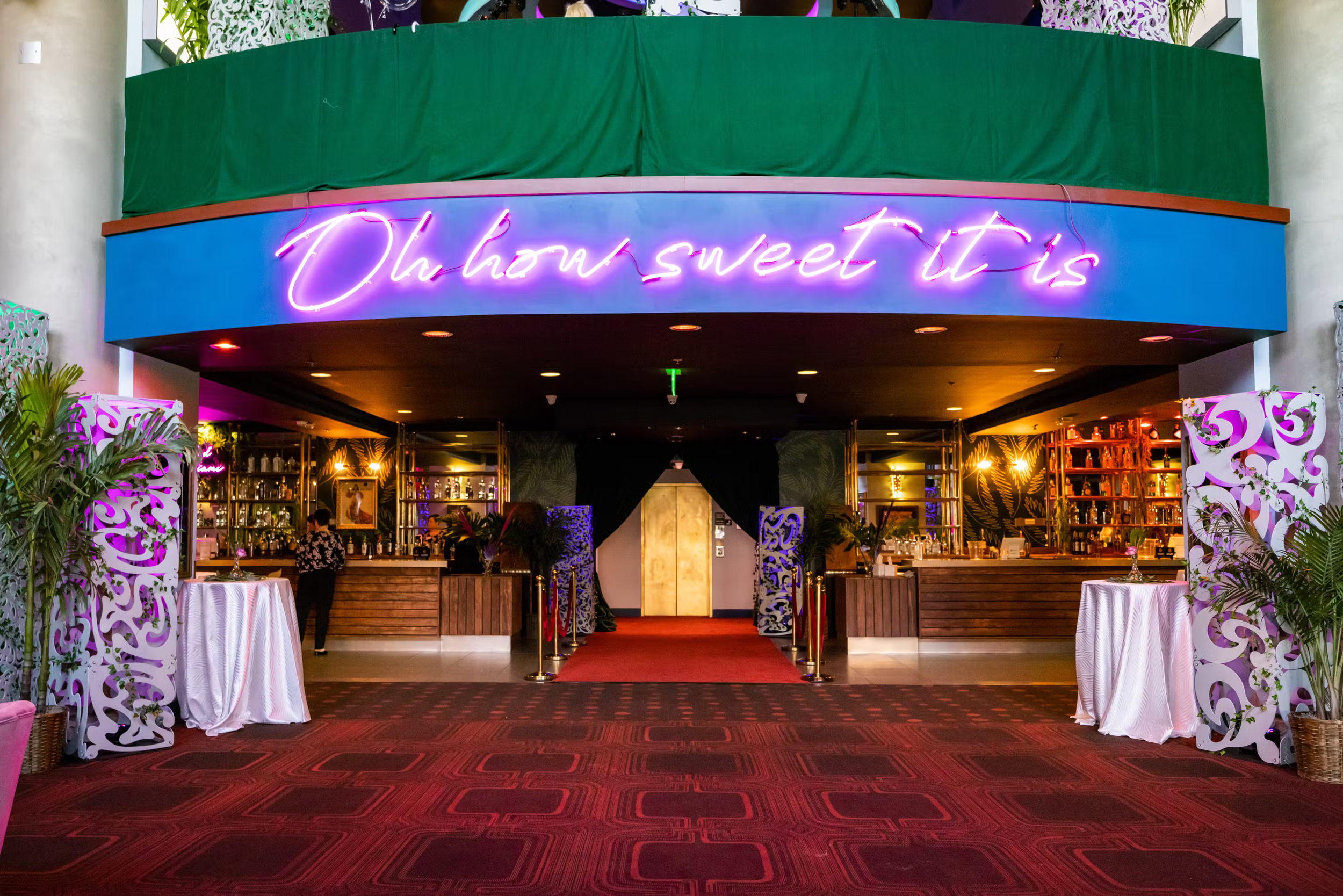 Entrance to a bar area with a neon sign that reads 'Oh how sweet it is'. The bar has purple and gold decor, with plants and white decorative panels on either side. There are red carpets and gold stanchions at the entrance to the bar.