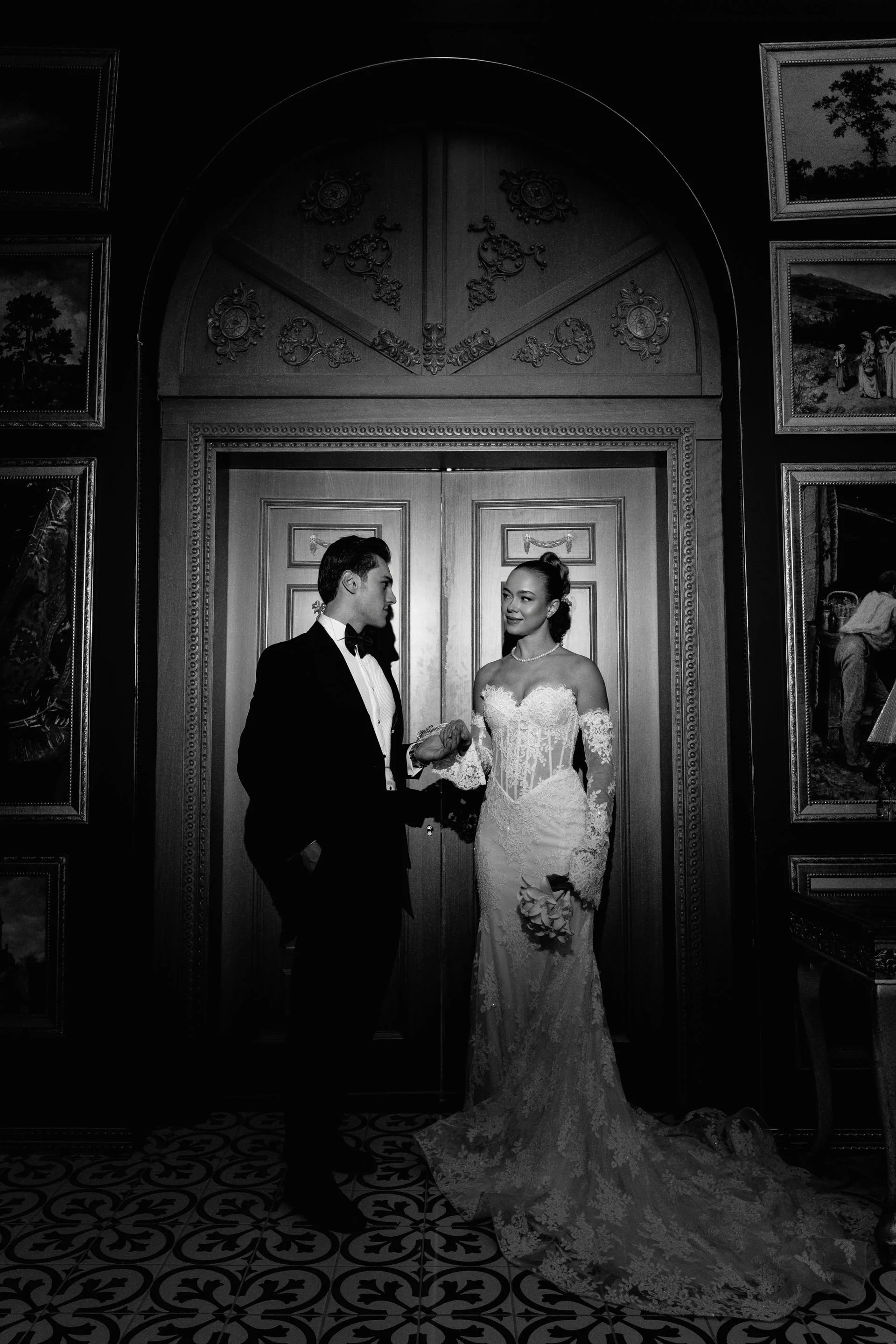 A black and white photo of a bride and groom holding hands and looking at each other, standing in front of ornate double doors with a decorative arch above, surrounded by framed landscape paintings on the walls.