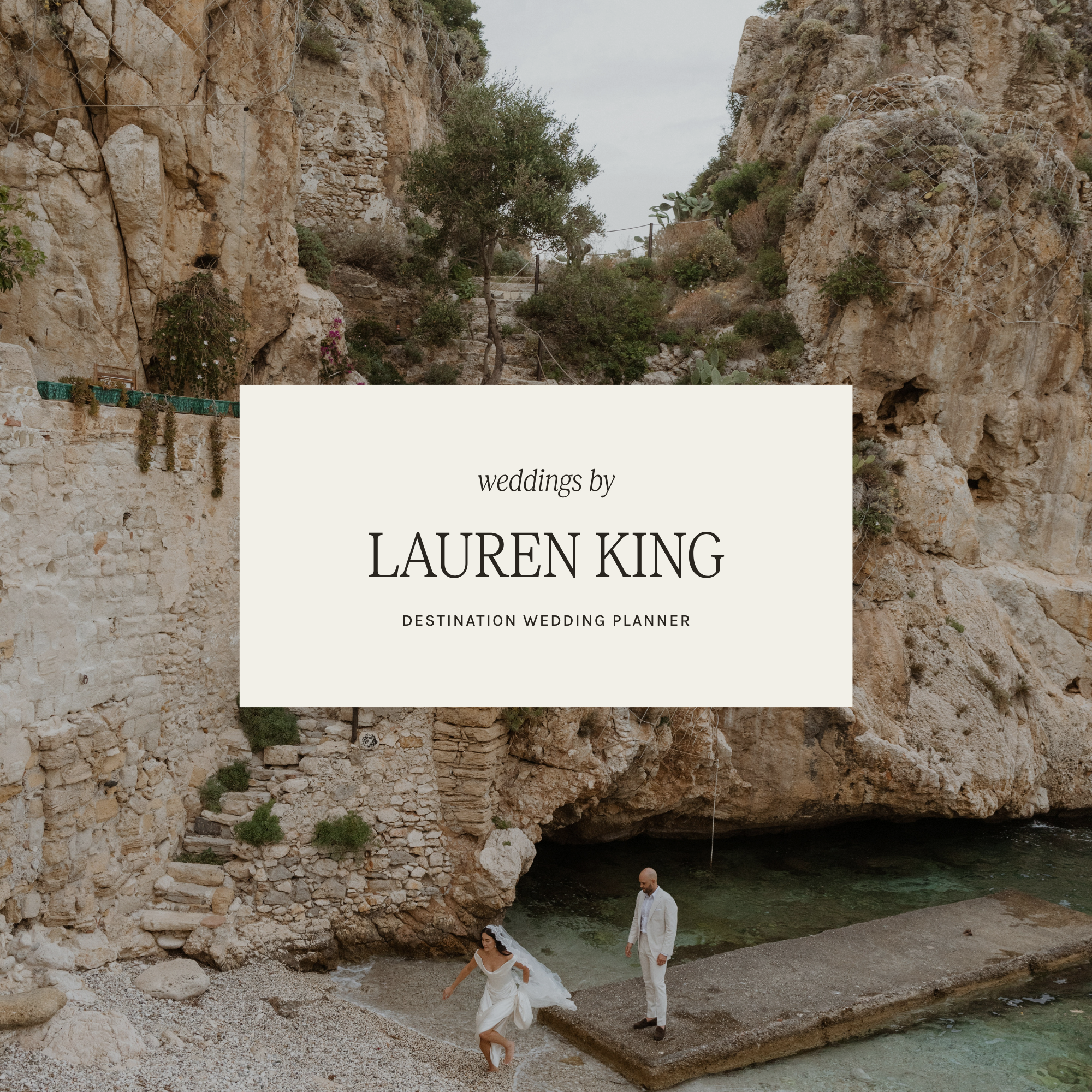 A bride in a white wedding dress and a groom in a white suit have a wedding ceremony by a river, with rocky cliffs and sparse greenery in the background.