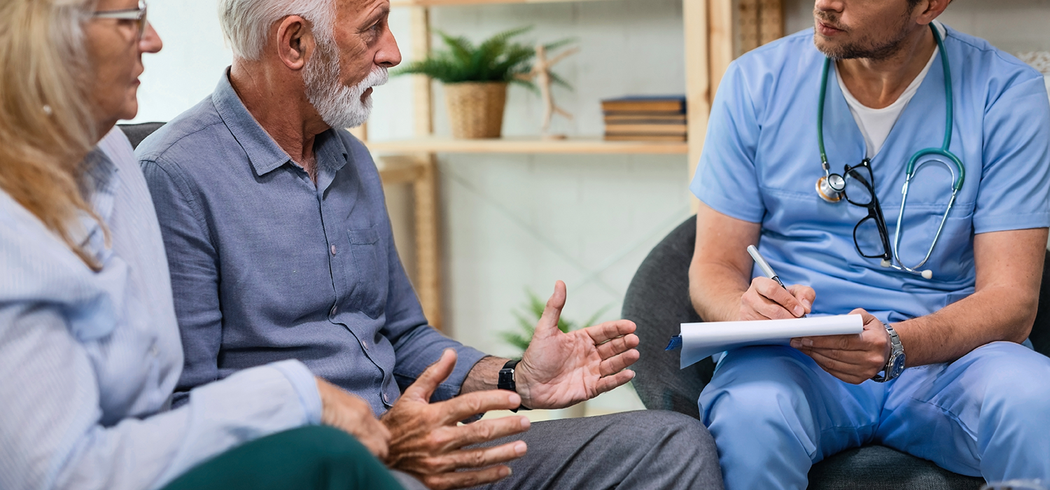 An elderly man in a blue shirt talking to a healthcare professional in blue scrubs, who is taking notes on a notepad. Two other women are partially visible, sitting beside the elderly man.
