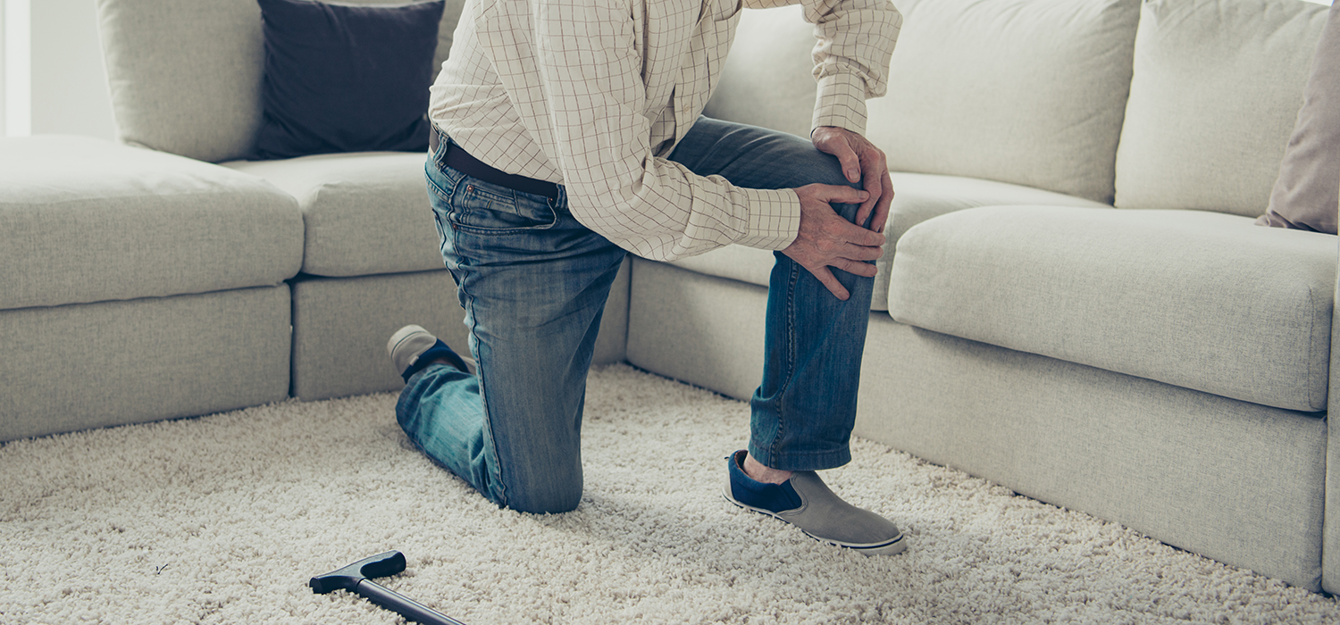 Man kneeling on a carpeted floor, holding his knee, suggesting knee pain, in a living room with a beige sofa and a black remote control nearby.