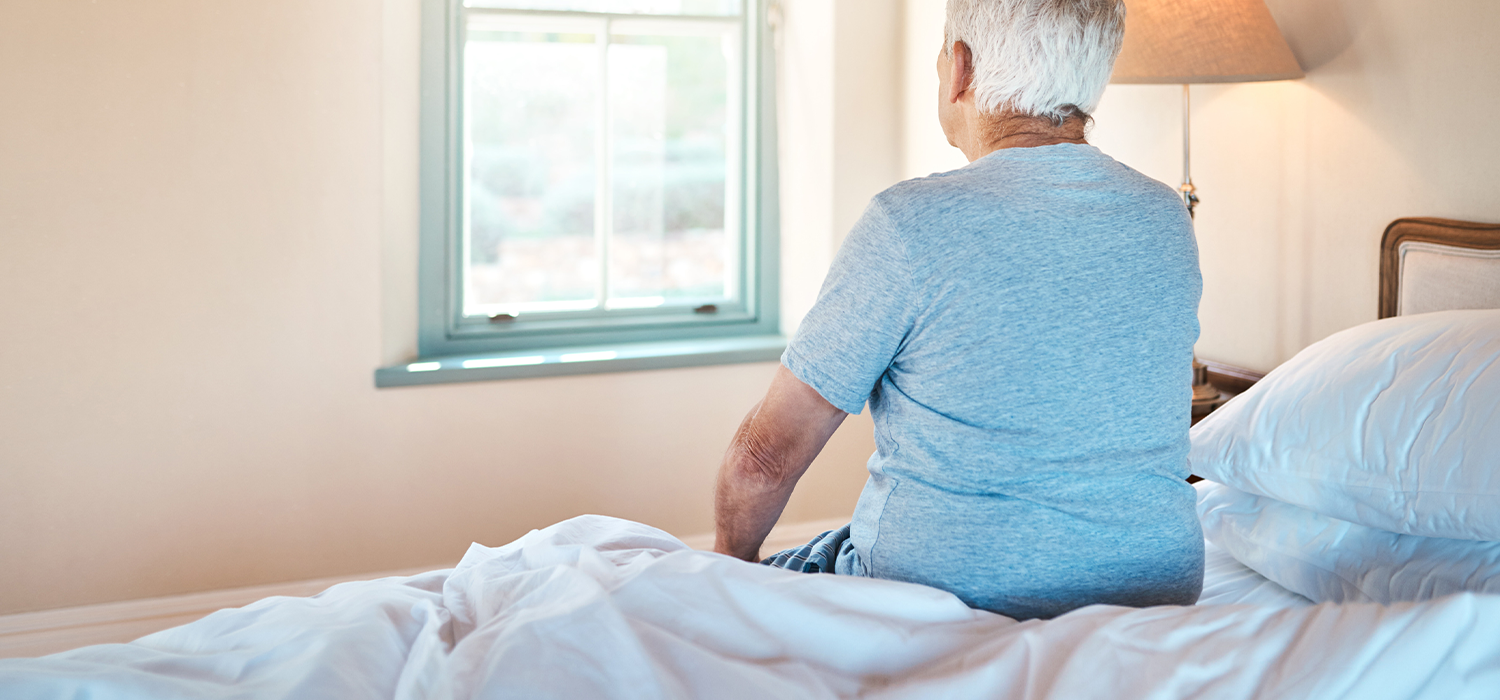 Older man with gray hair sitting on a bed in a bedroom, looking out the window with sunlight coming in, near a bedside lamp.