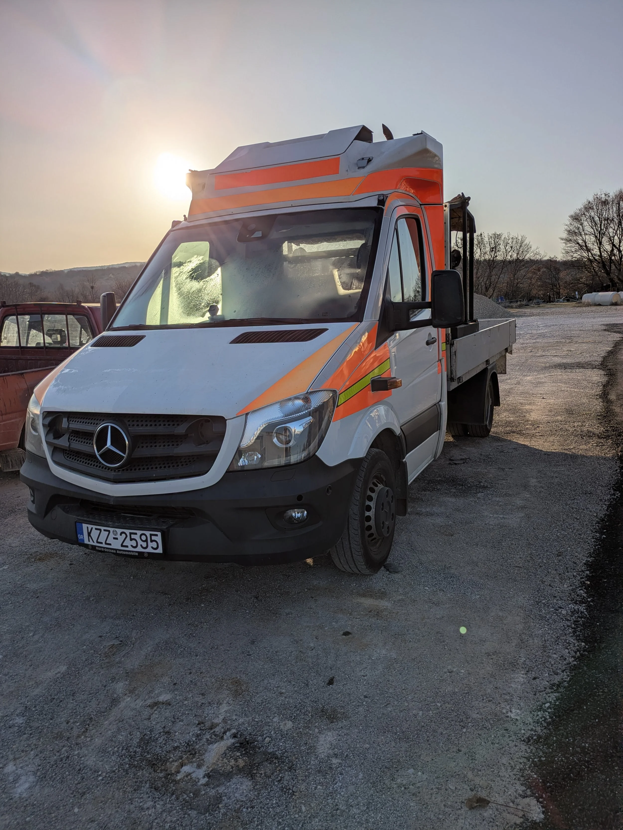 A white Mercedes-Benz utility truck with orange and yellow safety stripes parked outdoors at sunset, with a scenic background of hills and trees.