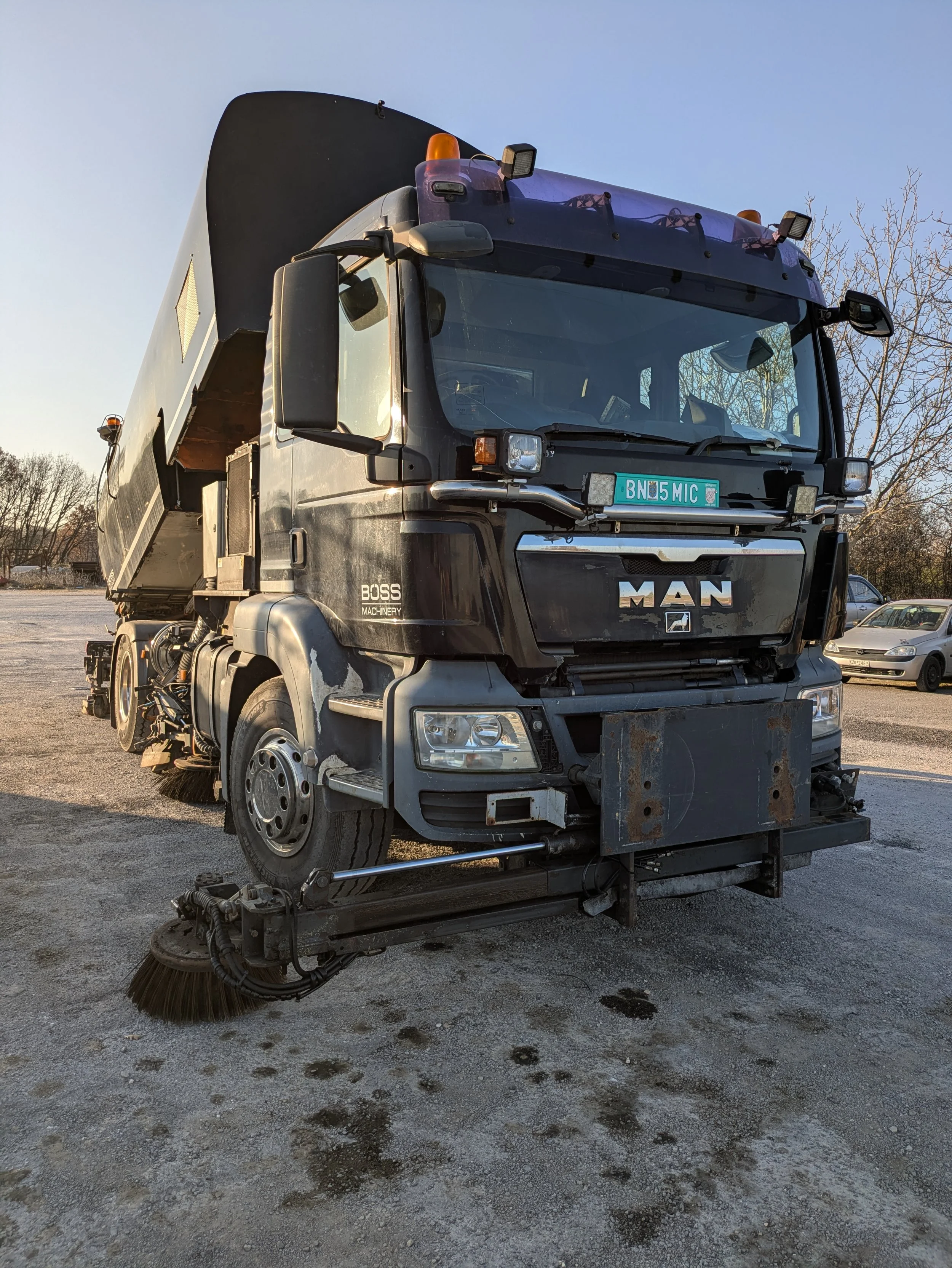 A black street sweeper truck parked on a gravel lot with trees and cars in the background.