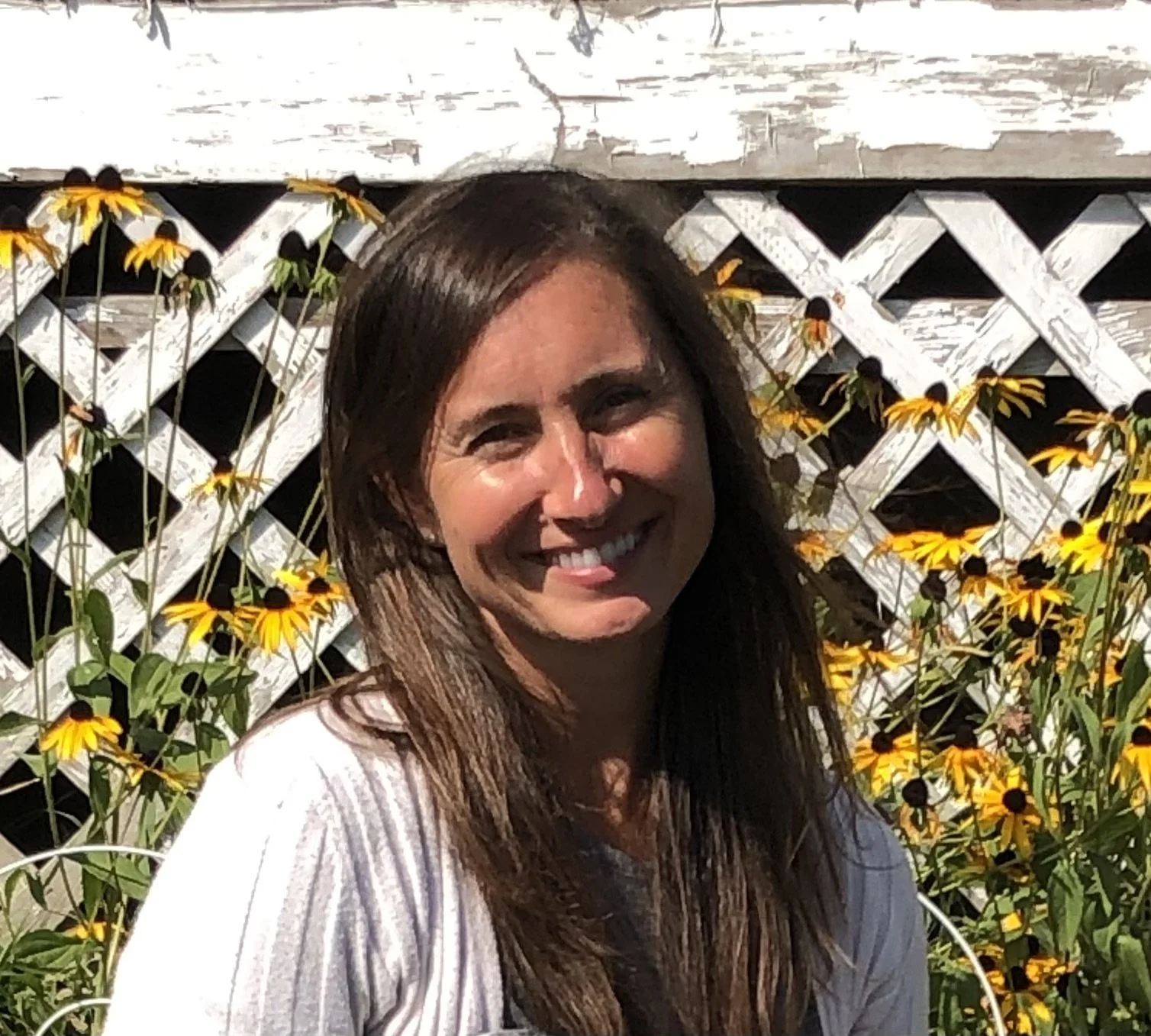 A woman with long brown hair smiling outdoors, standing in front of a white lattice fence decorated with yellow and black flowers.