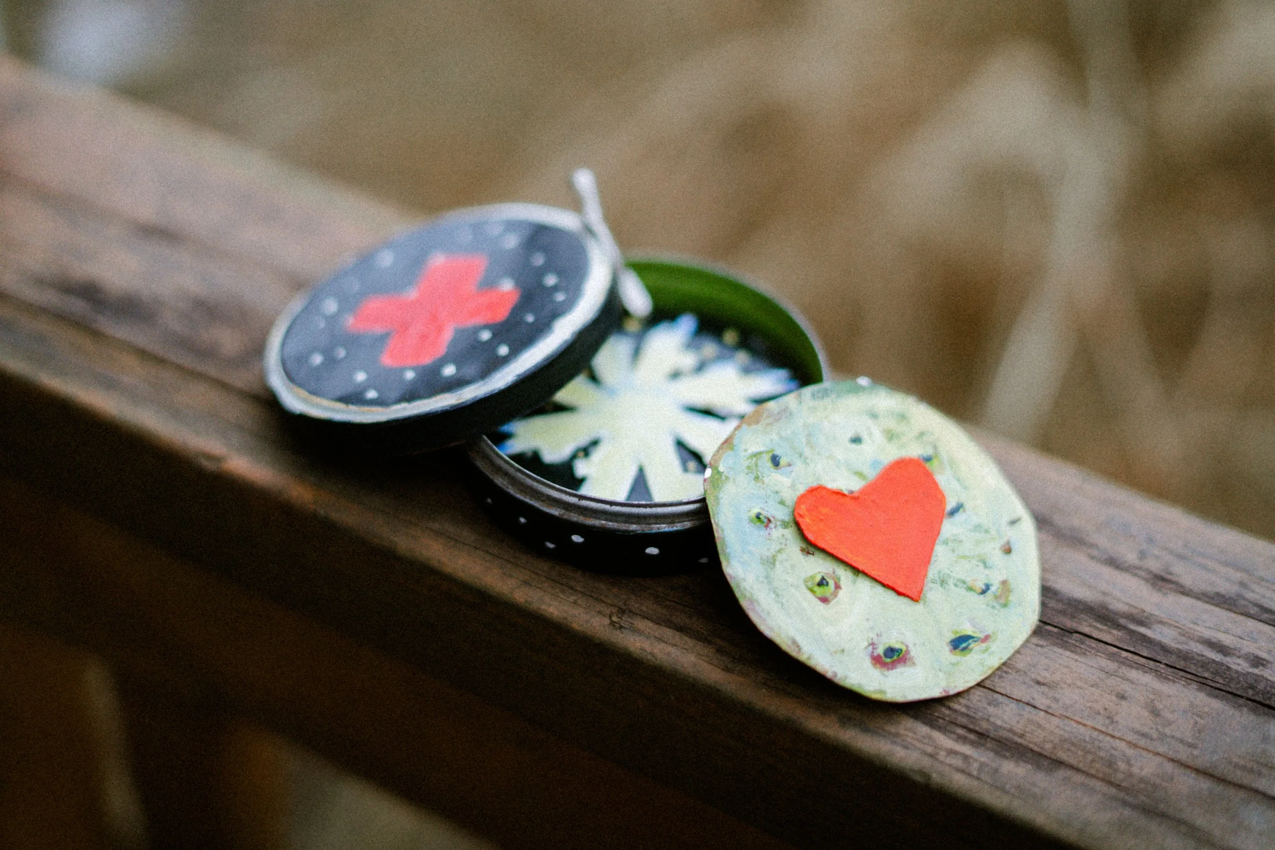 A round, decorated tin box with a slightly open lid, revealing a green interior with a snowflake design, sits on a wooden surface. The lid features a red heart on a green background with decorative peacock feathers, and the box's top layer has a black background with a red cross and white dots.