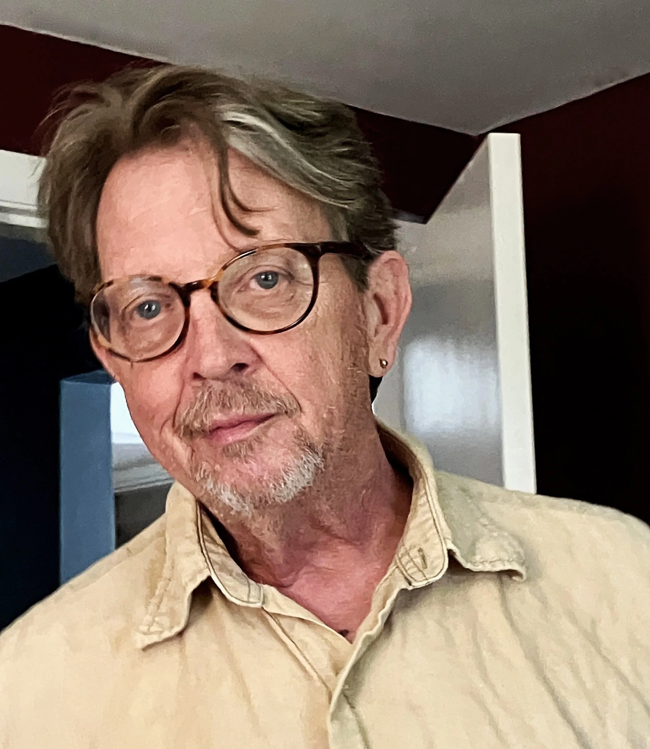 A man with glasses and gray hair, wearing a beige collared shirt, standing indoors in front of a doorway.